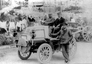 Woche von Nizza im März 1899. Wilhelm Bauer auf Daimler „Phönix“-Rennwagen gewinnt die Tourenfahrt Nizza–Magagnosc–Nizza über 85 Kilometer am 21. März 1899. Am Fahrzeug lehnt Hermann Braun, Chauffeur und Mechaniker von Emil Jellinek. Jellinek selbst nimmt auch an der Rennwoche teil - zum ersten Mal unter dem Pseudonym “Monsieur Mercédès”. 